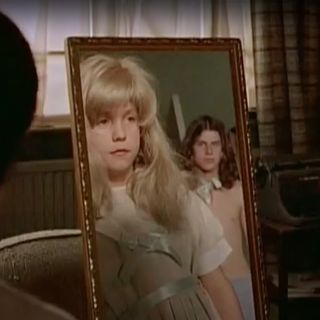 A young boy in a wig and a dress stares at his reflection in a mirror
