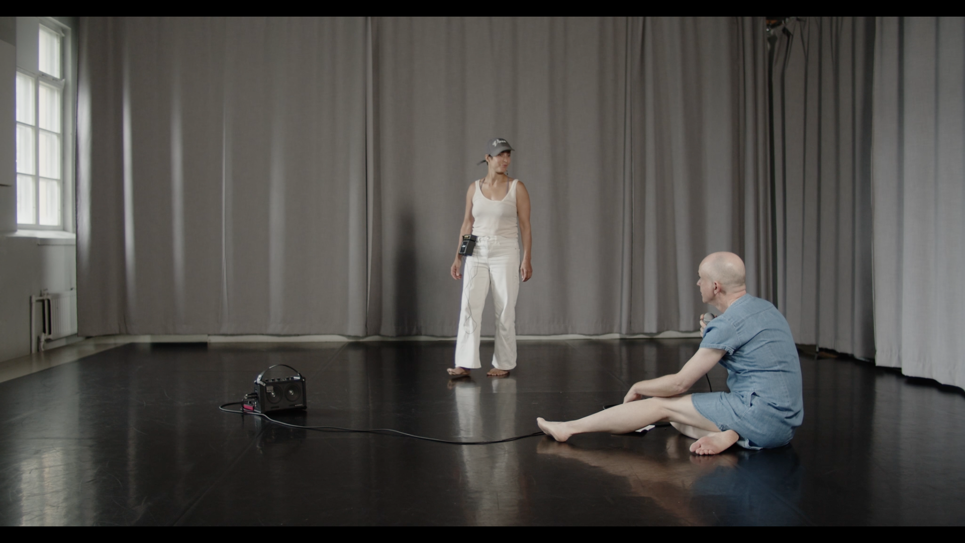 A trans woman in a denim dress sits on the floor in front of a queer woman